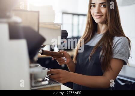 Portrait d'une jeune femme Barista utilisant une machine à café pour préparer des grains de café fraîchement moulus. Regarder l'appareil photo sourire. Banque D'Images