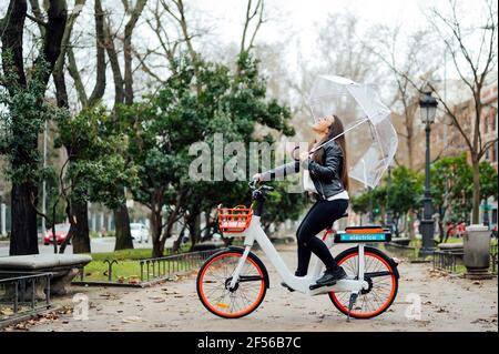 Femme insouciante assise sur un vélo électrique tout en tenant un parapluie dans la rue en ville pendant l'automne Banque D'Images