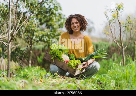 Femme souriante aux cheveux bouclés tenant une caisse de légumes en bois pendant s'accroupir dans le jardin Banque D'Images