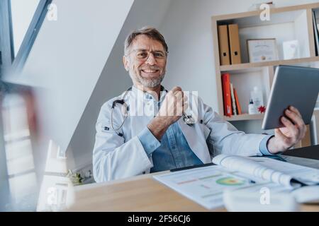 Un médecin souriant avec une tablette numérique, assis sur le bureau cabinet du médecin Banque D'Images