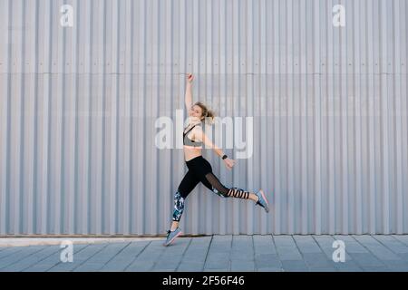 Femme souriante avec les bras relevés sautant sur le trottoir par le mur Banque D'Images