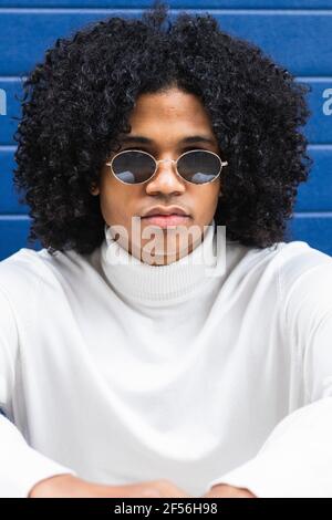 Cheveux bouclés adolescent garçon portant des lunettes de soleil assis contre le mur bleu Banque D'Images