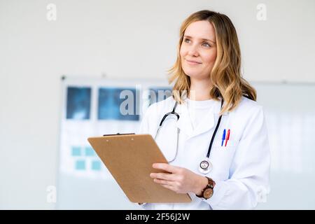 Femme blonde souriante médecin tenant le presse-papiers regardant loin Banque D'Images