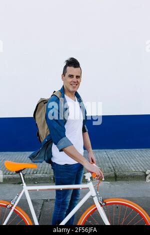 Homme souriant et charmant avec sac à dos tenant un vélo tout en marchant route Banque D'Images