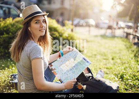 Une jeune femme voyageant à l'exploration de la carte et de la ville écoute de la musique en streaming dans des écouteurs, regardant l'appareil photo sourire. Banque D'Images