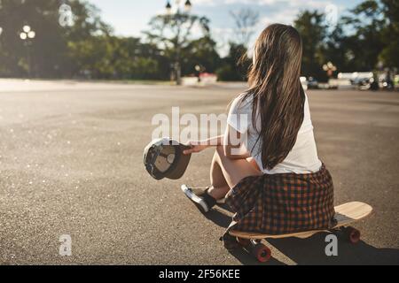 Loisirs, passe-temps et skate dans le concept de la ville. Vue de l'arrière à emporter par une adolescente vivane admirez le parc à roulettes vide et le soleil du soir en vous asseyant Banque D'Images