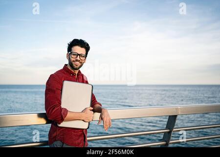 Homme d'affaires souriant avec un ordinateur portable penché sur la rampe au bord de la mer Banque D'Images