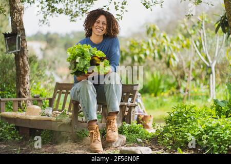 Femme souriante tenant des légumes feuillus tout en étant assise sur un banc en bois dans le jardin Banque D'Images