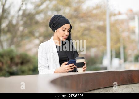Jeune femme arabe utilisant un smartphone tenant une tasse réutilisable pendant se tenir au mur de retenue Banque D'Images