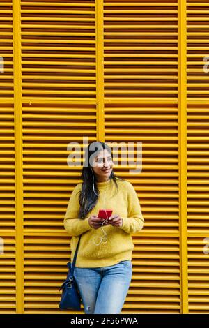 Femme souriante avec téléphone portable qui écoute de la musique avec un casque en vous tenant contre le mur jaune Banque D'Images