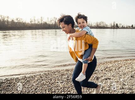 Une femme gaie qui se fait dormer avec un garçon souriant tout en courant sur le bord du lac jour ensoleillé Banque D'Images