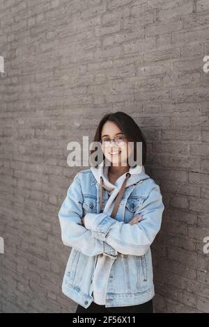 Portrait vertical d'une étudiante féminine intelligente et attirante, des femmes en 20 ans, debout sur un mur de briques à l'extérieur, appareil photo souriant satisfait et lui-même Banque D'Images