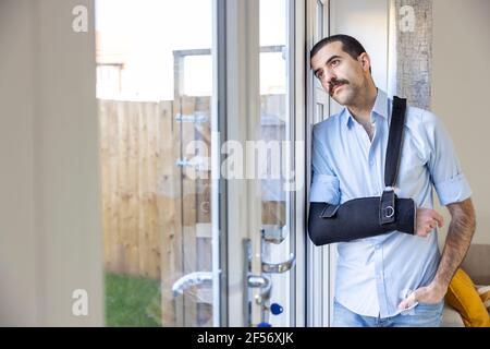 Homme blessé portant une élingue de bras debout tout en regardant par la fenêtre Banque D'Images