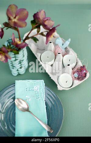 Photo de vase avec roses de Noël (Helleborus niger), serviette verte sur plaque verte et carton d'œufs avec bougies placées à l'intérieur des coquilles d'œufs Banque D'Images