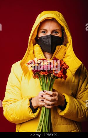 Femme portant un masque facial et une chemise à capuchon tenant la fleur pendant sur fond rouge Banque D'Images