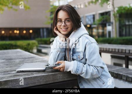 Portrait en gros plan d'une jeune étudiante gaie avec des cheveux courts, se pencher sur la paume en regardant mignon l'appareil photo avec un sourire heureux, assis près de l'ordinateur Banque D'Images