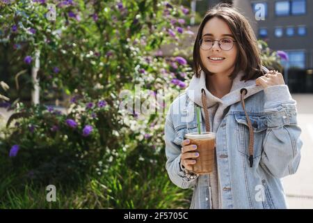 Portrait d'une jeune femme attrayante debout dans la rue près des buissons verts, boire un café préféré, glacière à emporter un café local impressionnant, souriant Banque D'Images