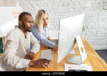 Jeunes collègues adultes concentrés assis à un bureau, utilisant un ordinateur, travaillant ensemble. Des partenaires d'affaires sérieux à la recherche d'idées de projet Banque D'Images