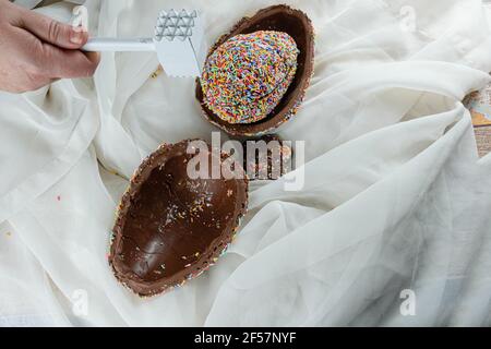 Main de femme avec un marteau sur plusieurs moitiés de l'oeuf de Pâques (pinata). Œuf recouvert de saupoudrer de chocolat coloré. Banque D'Images