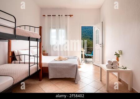 Intérieur blanc moderne de la chambre familiale avec lit double classique en bois et lits superposés avec escalier en métal, porte ouverte sur l'été vue sur le jardin d'oliviers, balcon, Banque D'Images