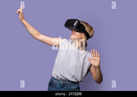 La femme caucasienne aux cheveux blonds a la première réalité virtuelle touchez quelque chose en portant un casque sur un violet mur du studio Banque D'Images