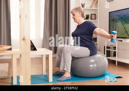 Grand-mère faisant du sport en pyjama avec entraîneur personnel en ligne. Personne âgée pensionné en ligne entraînement à l'exercice sur Internet à la maison activité sportive avec haltères, bande de résistance, ballon suisse à l'âge de la retraite Banque D'Images