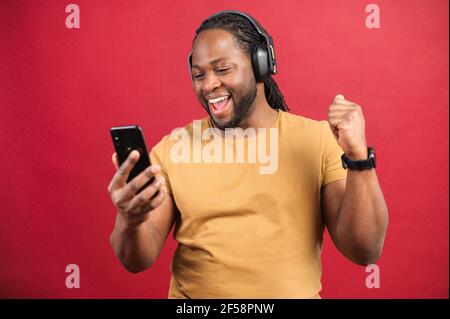 Portrait studio de jeune Afro-américain avec des dreadlocks dans des écouteurs sans fil isolés sur le mur rouge disant oui, regardant le téléphone mobile, appréciant l'écoute de la bande-son musicale préférée, dansant Banque D'Images