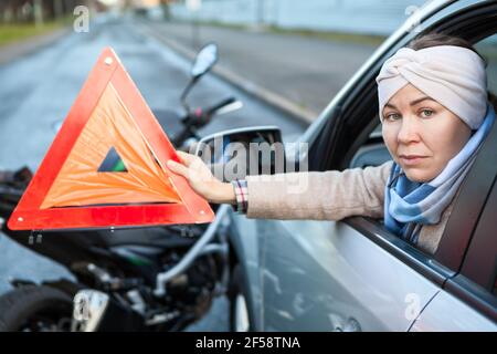 Femme un conducteur montrant un triangle de signalisation d'urgence en étant assis dans une voiture après un accident de la route avec moto, ne quitte pas la voiture Banque D'Images