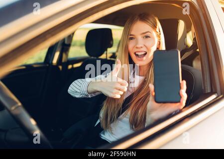 Jeune femme heureuse conduisant une voiture, elle montre un écran de téléphone vide et un pouce vers le haut, applications mobiles utiles pour un conducteur Banque D'Images