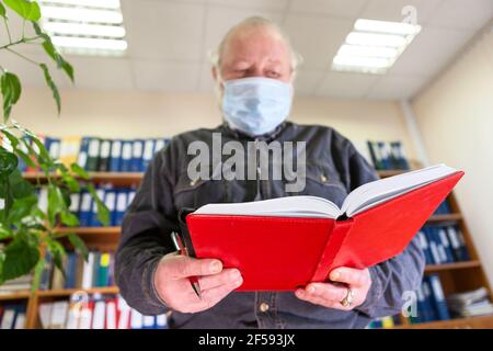 Archiviste mâle mature tenant un cahier ouvert dans les mains, regardant la caméra, homme caucasien portant un masque facial en raison de la pandémie Covid-19. L'accent est mis sur les organi rouges Banque D'Images