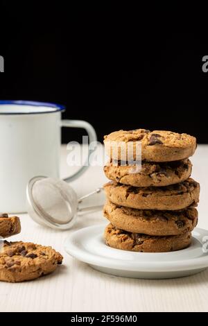 Gros plan d'une pile de biscuits au chocolat dans une assiette blanche et une tasse blanche, sur une table en bois blanc, sur fond noir, à la verticale, avec espace de copie Banque D'Images