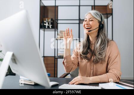 Confiante et influente joyeuse femme d'affaires asiatique mûre aux cheveux gris, téléphoniste de centre d'appels, discutant avec des collègues ou des amis pendant un appel de vidéo conférence en ligne, salue, souriant amical Banque D'Images
