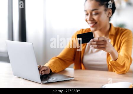 Une jeune fille afro-américaine souriante et défochée, vêchée de vêtements élégants et décontractés, assise à la maison, tient une carte bancaire, utilise un ordinateur portable, effectue un paiement en ligne pour des achats ou des factures, saisit un numéro de carte Banque D'Images