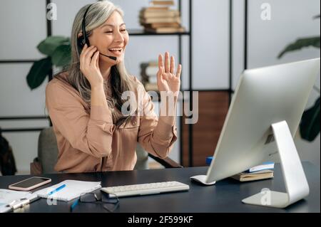 Femme d'affaires asiatique, responsable, coach, portant un casque, chat avec des partenaires d'affaires par conférence téléphonique, briefing en ligne, gestuelle, sourire amical Banque D'Images