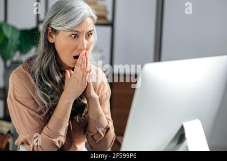Stupéfait femme d'affaires asiatique d'âge moyen aux cheveux gris, gestionnaire principal, courtier immobilier, s'asseoir à table et recevoir des nouvelles inattendues, message ou rapport financier, regardant l'écran avec une expression choquée Banque D'Images