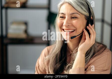 Gros plan sur une femme asiatique mûre et mûre aux cheveux gris joyeuse, employée d'un centre d'appels, communique avec ses collègues ou ses clients par vidéoconférence, consultation en ligne, utilisation d'un micro-casque, sourire Banque D'Images