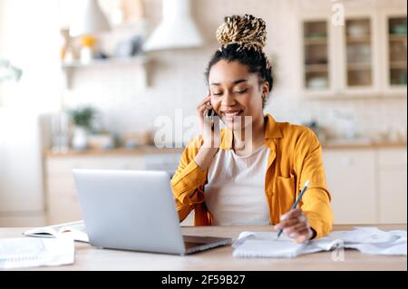 Jeune femme afro-américaine habillée avec élégance, femme d'affaires moderne ou agent immobilier, ayant une conversation téléphonique agréable avec un client ou un employé, assis sur le lieu de travail, souriant