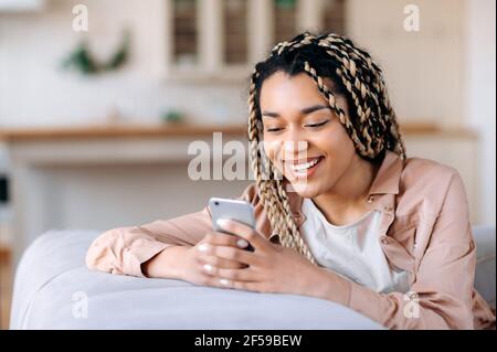 Une jeune fille afro-américaine attirante et satisfaite est assise dans le canapé, utilise un smartphone, passe du temps sur les réseaux sociaux, a reçu de bonnes nouvelles ou messages, navigue sur Internet, discute en ligne avec ses amis ou sa famille Banque D'Images