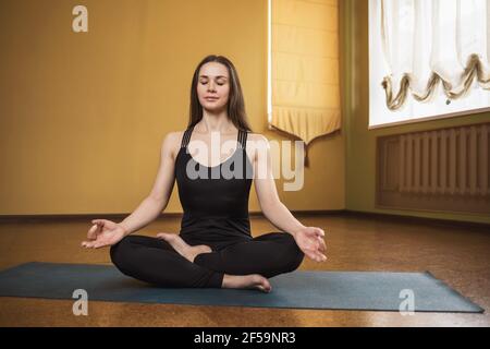 Jeune femme attirante en noir sportswear, pratiquant le yoga, est engagée dans la méditation dans la position lotus sur un tapis de gymnastique dans le studio. Banque D'Images