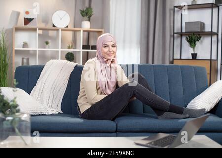 Portrait d'une femme arabe très agréable dans le hijab assise sur un canapé bleu dans le salon de la maison, penchée la tête à la main et regardant l'appareil photo avec le sourire. Banque D'Images