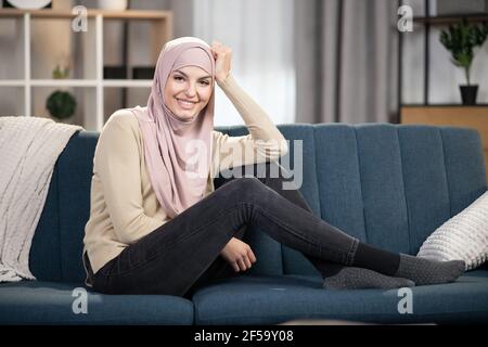 Portrait d'une femme musulmane gaie dans le hijab posant sur un canapé à la maison, se détendant dans le salon, appréciant le week-end ou le temps libre Banque D'Images