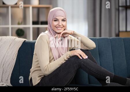 Portrait en gros plan d'une femme musulmane heureuse et détendue de 30 ans dans le hijab, assise sur le canapé bleu doux dans le salon confortable de la maison et regardant l'appareil photo avec le sourire. Banque D'Images