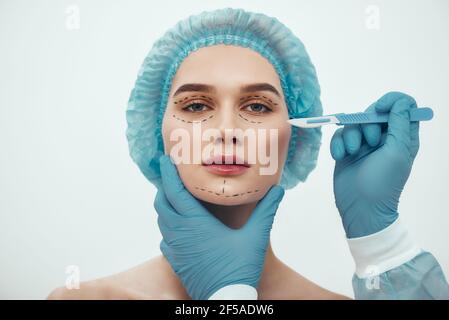 Chirurgie de levage de visage. Portrait d'une belle jeune femme en chapeau médical bleu ayant une chirurgie esthétique du visage. Chirurgien plastique en gants bleus tenant le scalpel. Banque D'Images