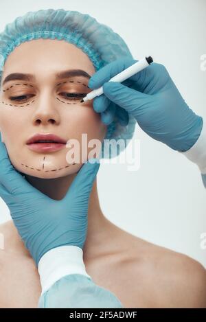 Beau patient. Jeune femme en chapeau médical bleu attendant la chirurgie du visage tandis que chirurgien en plastique en gants bleus dessinant des lignes pointillées sur son visage Banque D'Images