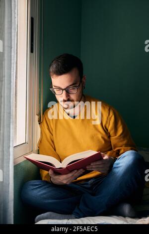 Un jeune homme lit un livre à côté de sa chambre fenêtre Banque D'Images