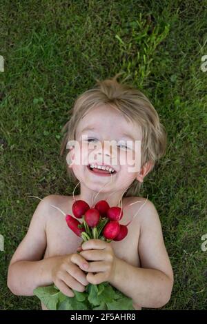 joyeux petit garçon mignon, allongé sur son dos sur de l'herbe verte, tenant dans les mains récolte fraîche de radis du jardin. peu d'aide, l'agriculture, saine eati Banque D'Images