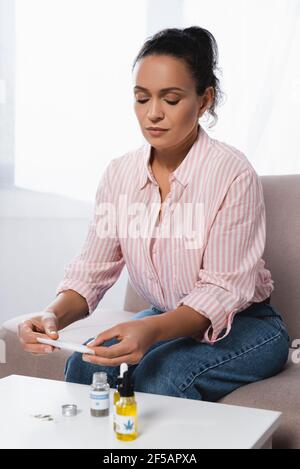 femme afro-américaine assise sur un canapé tout en roulant avec du papier cannabis médical séché près des bouteilles de cbd Banque D'Images