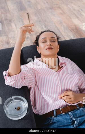vue en grand angle sur une femme afro-américaine posée sur un canapé et fumer roulé joint avec du cannabis médical près du cendrier Banque D'Images