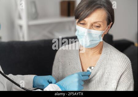 L'infirmière soignante d'une patiente âgée portant un masque à la maison pendant une pandémie de coronavirus, écoute avec un stéthoscope à la fréquence cardiaque et à la fréquence respiratoire. Rendez-vous du médecin à la maison Banque D'Images