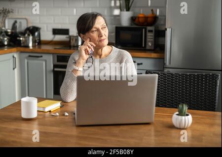 Femme pensive à la retraite utilisant un ordinateur portable pour le travail à distance de la maison, la femme senior s'assoit avec un ordinateur au bureau de cuisine, regarde loin et pense Banque D'Images